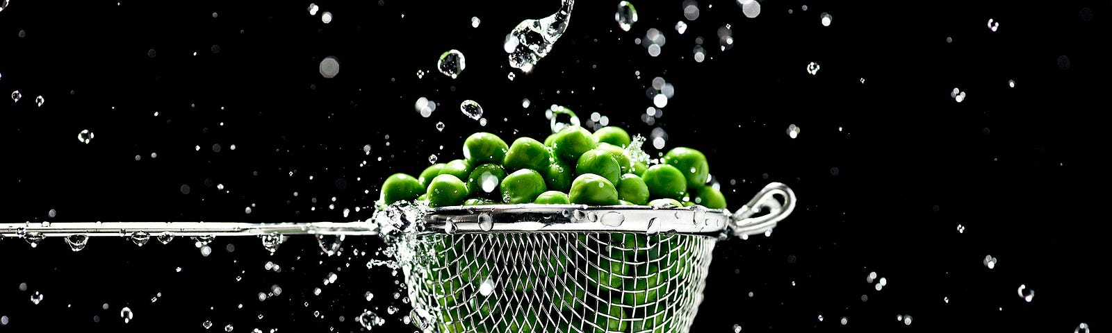 Strainer of fresh green peas and water droplets against black background.