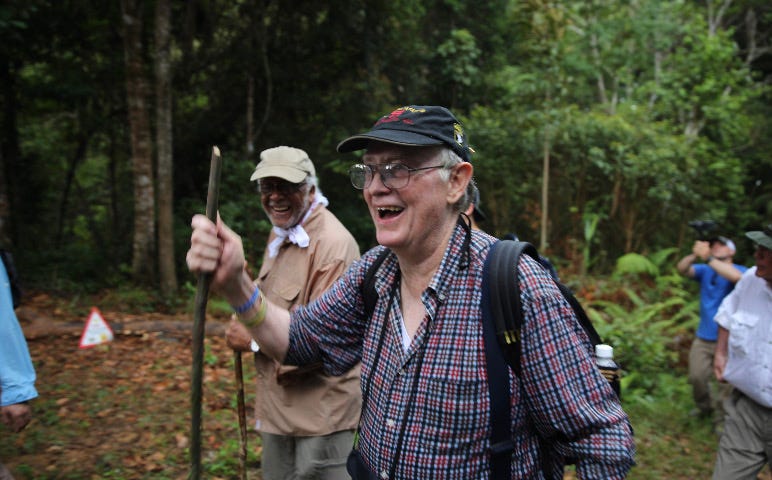 Two smiling men walking through the jungle with walking sticks.