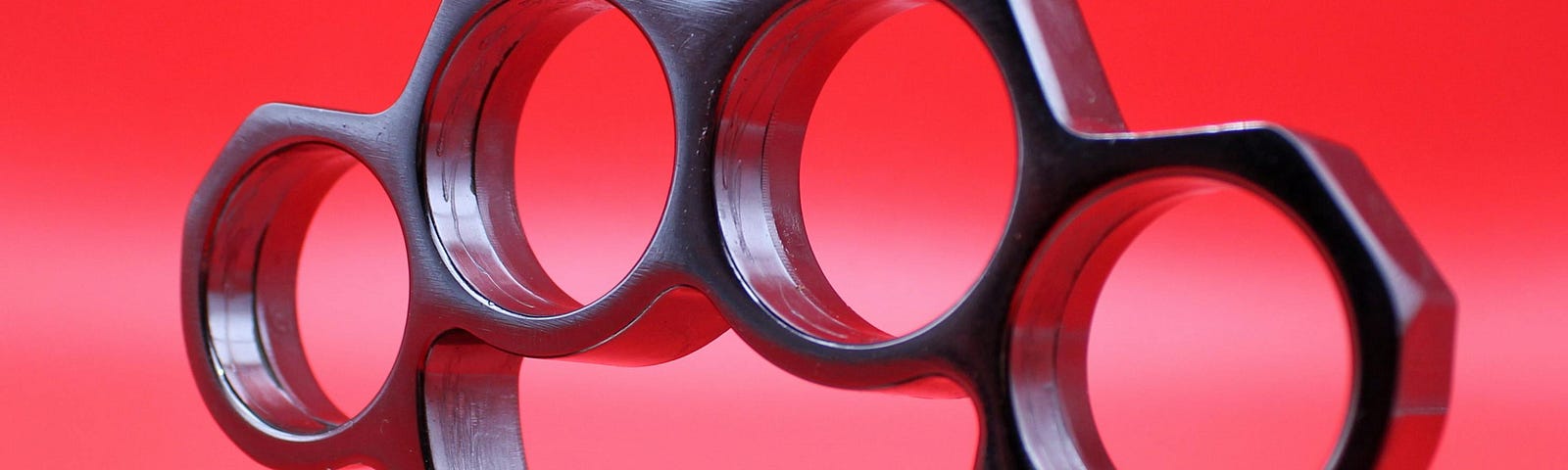 Image shows close-up of shiny, silver set of brass knuckles against a reddish pink background
