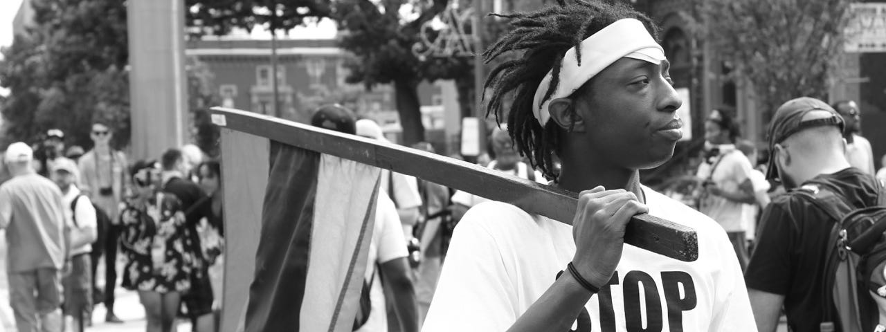 Black and White photo of a man holding a flag and wearing a shirt that states “Stop Killing Black People”