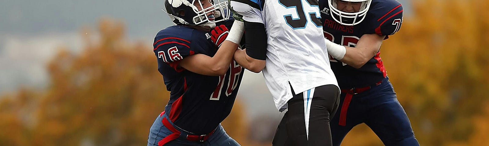 Three football players during a running play, two in dark blue uniforms and one in a white uniform.