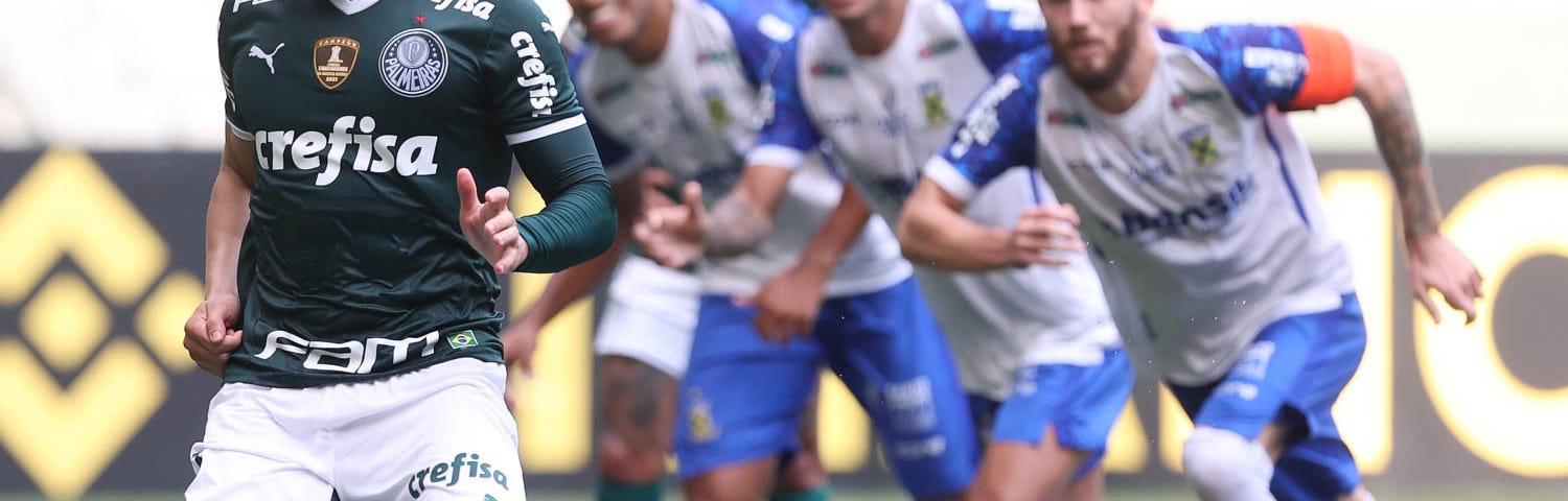 Raphael Veiga cobra pênalti com o pé esquerdo. Ele veste uniforme do Palmeiras: camisa e meias verdes, calção branco. A camisa é de marga curta Ele usa ainda um manguito verde no braço esquerdo. Atrás dele, três jogadores do Santo André, de camisa e meias brancas e calção azul, ameaçam invadir a área.