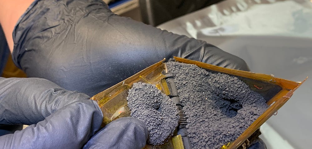 A hand holding a small tetrahedral robot, the other hand holding the sample chamber open to reveal it filled with fine-grained loose rocky material.