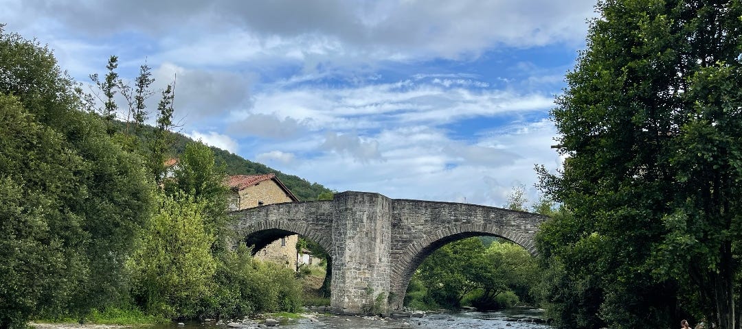 An ancient stone bridge with high arches over a tranquil river that you see in the foreground. It’s a sunny day, a blue sky with some smaller clouds. Left and right of the river are green trees.