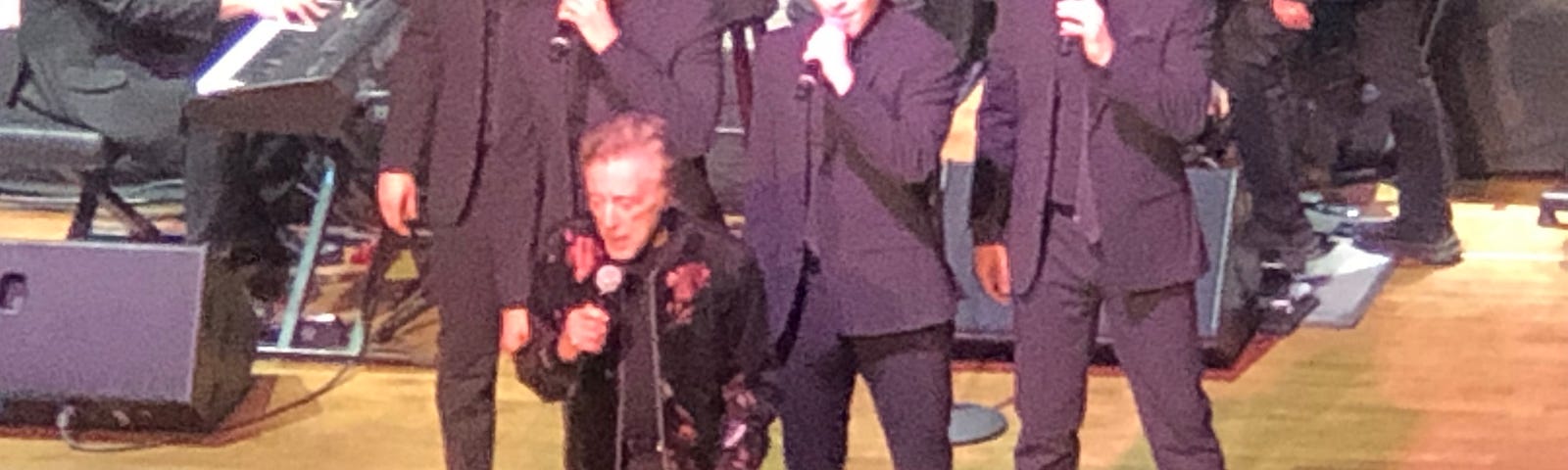 Photo of Frankie Valli and The Four Seasons. Frankie Valli is in the front, with The Four Seasons behind him. All are singing into microphones they hold in their hands.