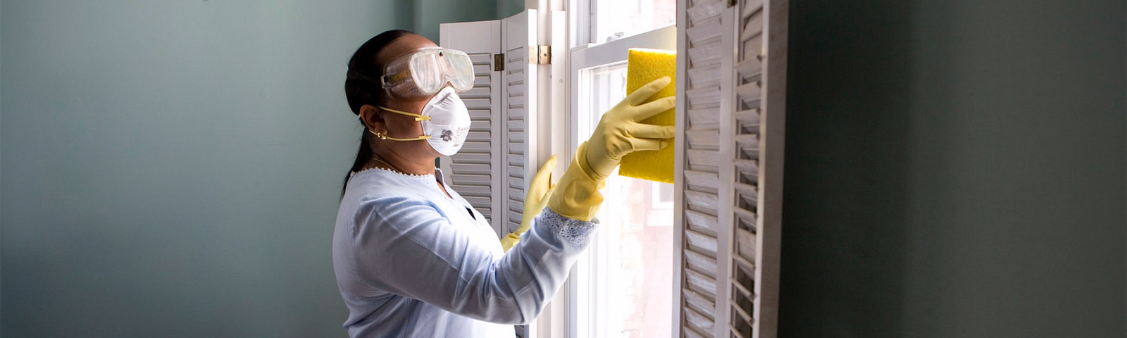 a woman with goggles, mask, and yellow gloves cleans a window with a sponge