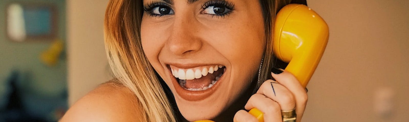 A bright-eyed young woman with blonde hair, wearing a floral brown dress and a big smile is speaking into a yellow handset telephone receiver.