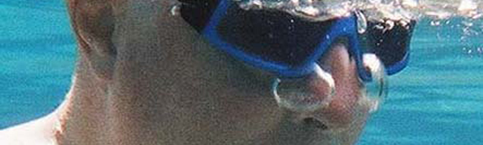 An older man under water in a pool exhales 2 bubbles through his nose.