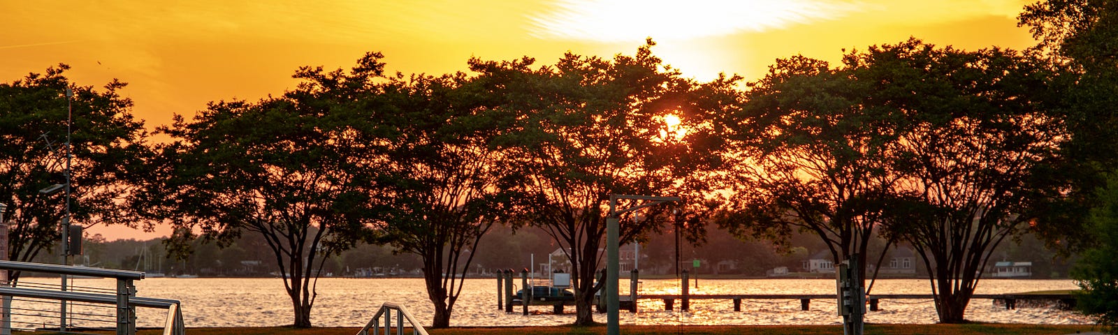 An orange sky over a lake, a park and a wooden bridge in the foreground, and the sun partially hidden behind the trees