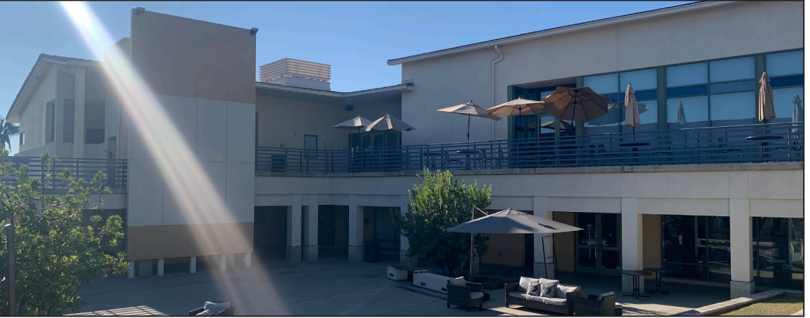 An image of Whittier College’s Campus Courtyard. Tables with umbrellas are displayed on the second level while a couch with a single large umbrella is above it on the lower level. There is a sun glare on the left side of the photo.