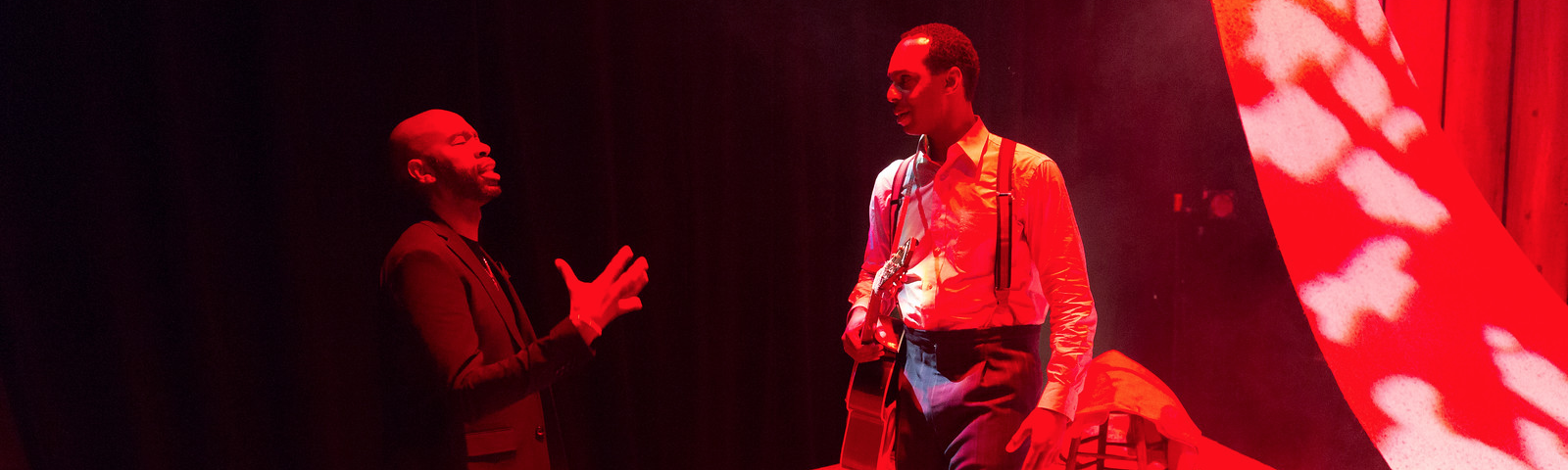 A man in a black suit and a man in a white shirt, suspenders, and black pants holding a guitar talk onstage in red light.