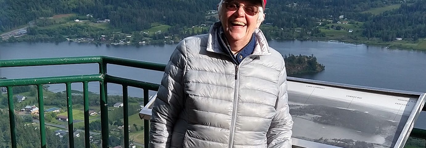 Picture of the author in a red cap and light grey jacket standing in front of a lookout with her dog K.C. a miniature Schnauzer. The scene behind is a beautiful vista of islands dotting the water below.