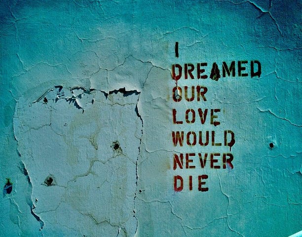 Text: “I Dreamed Our Love Would Never Die” stenciled on a faded, water stained, and peeling blue wall.