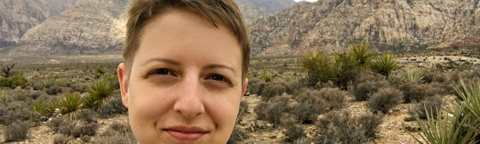 Selfie photograph of a white woman with short brown hair standing in the desert. She is slightly smiling at the camera. There are rock formations in the distance and shrubby plants in the foreground.