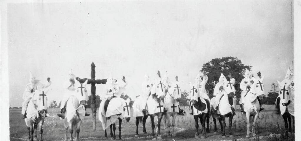 Nine hooded members of the KKK on horseback, with a cross in the background.