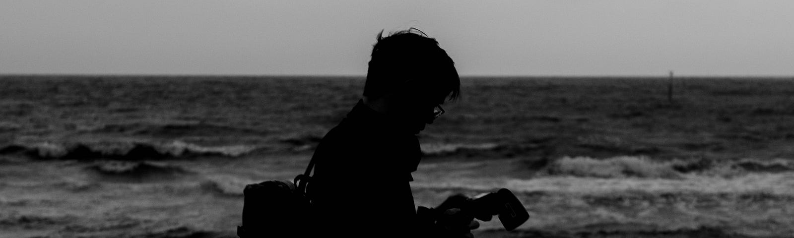 A black-and-white photo from Hanying Xie’s photobook, Behind the Camera. Photo of a man with a camera, silhouetted against a beach. Waves cascade onto the sand behind him. He stares down at his camera’s screen, contemplating his shots.