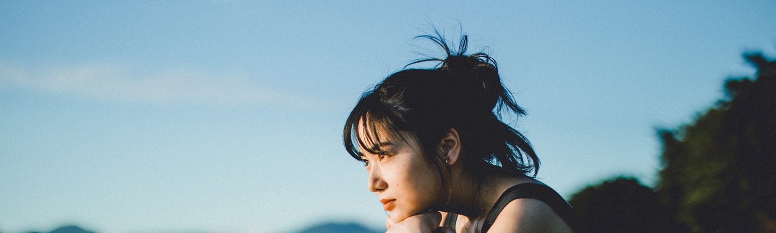 A sad (but hopeful) woman with dark hair sits on a beach and stares over the water.