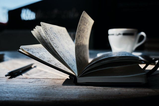 An open book sits on a table, with pages standing up, others falling down, pen writing is visible on the pages. Mug of coffee or tea in the background, a pen sits near the book.