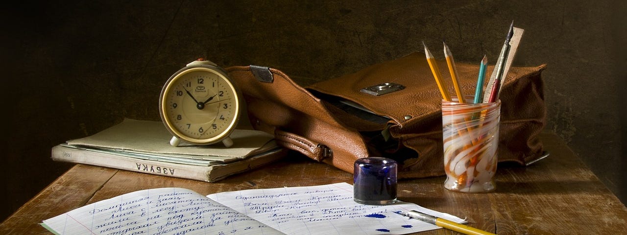 A writer’s desk with an open notebook, inkwell and stylus, ink drops over written words. Satchel, analog clock and pencil cup accompany notebook.