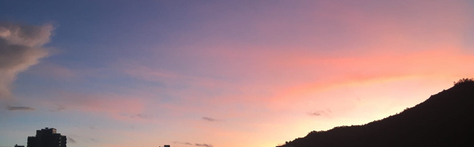 The pastel-tinted sky of sunrise with the black silhouette of mountains & buildings in front.