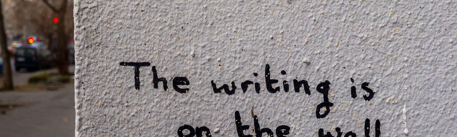 Muro branco com texto em inglês escrito “The writing is on the wall”