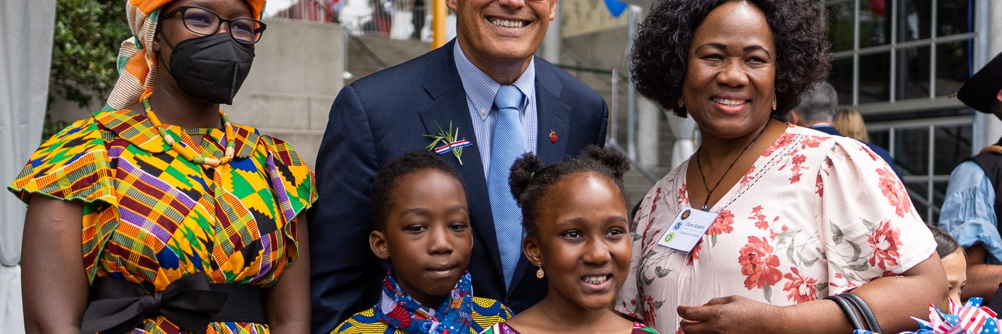 Gov. Jay Inslee poses for a photo with two women and two young girls, who are all dressed in bright clothing and smiling at the camera. One young girl is holding a small American flag.