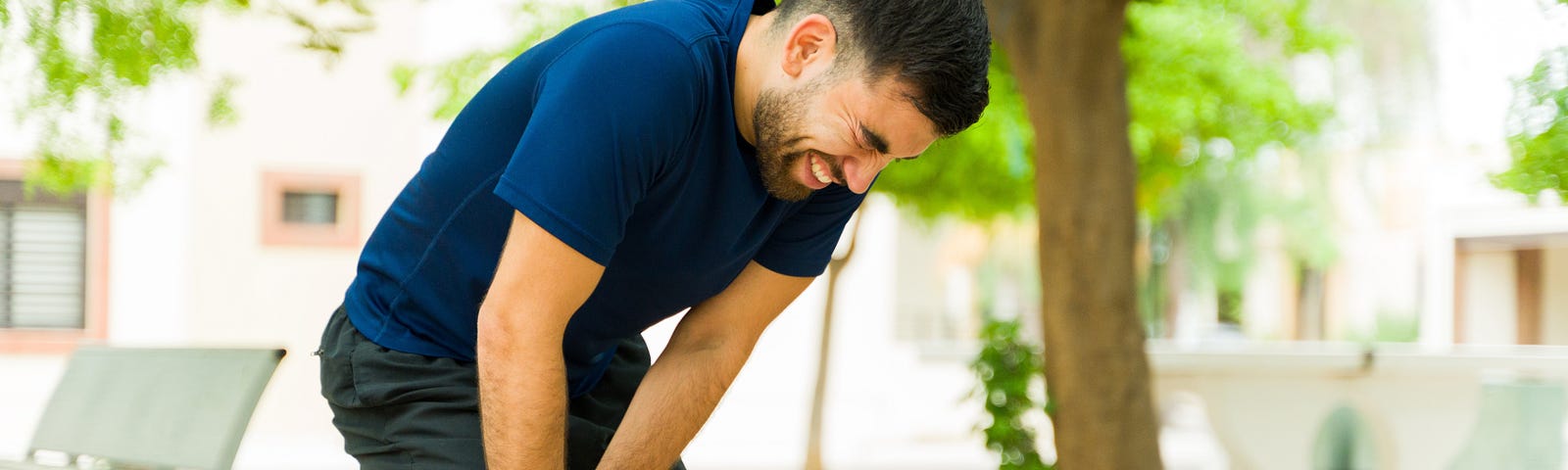 Man in running clothes grimacing in pain, clutching his knee outside in a park