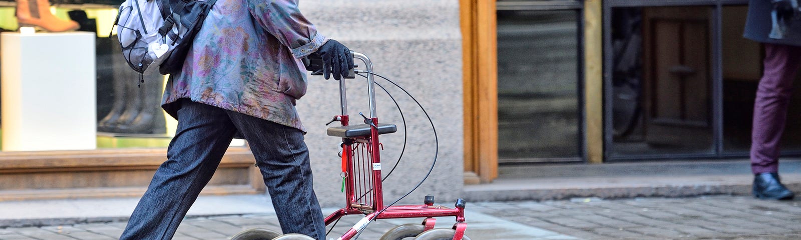 An older woman walking with the support of a rollator on the sidewalk.