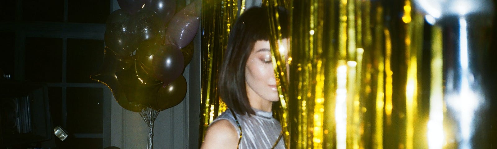 A young, thin, attractive woman who shoulder length black hair and a party dress is half hidden behind a gold foil curtain. Another woman and balloons, in less light, are in the background.