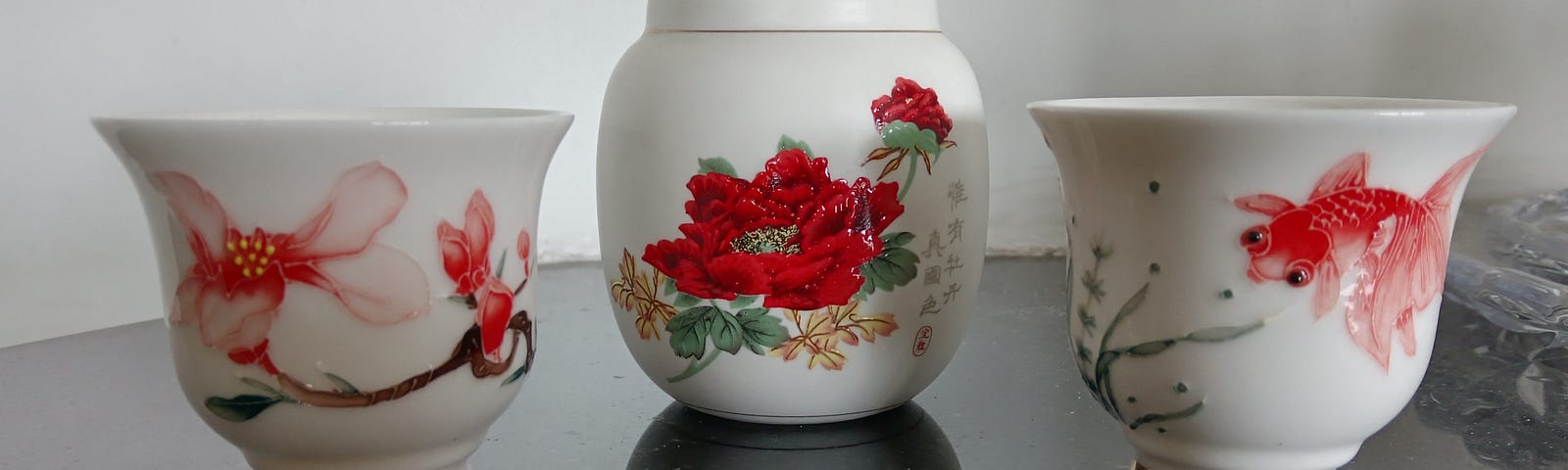 2 Fine China tea cups embellished with a flower and a goldfish beside a flowered Chinese tea caddy.