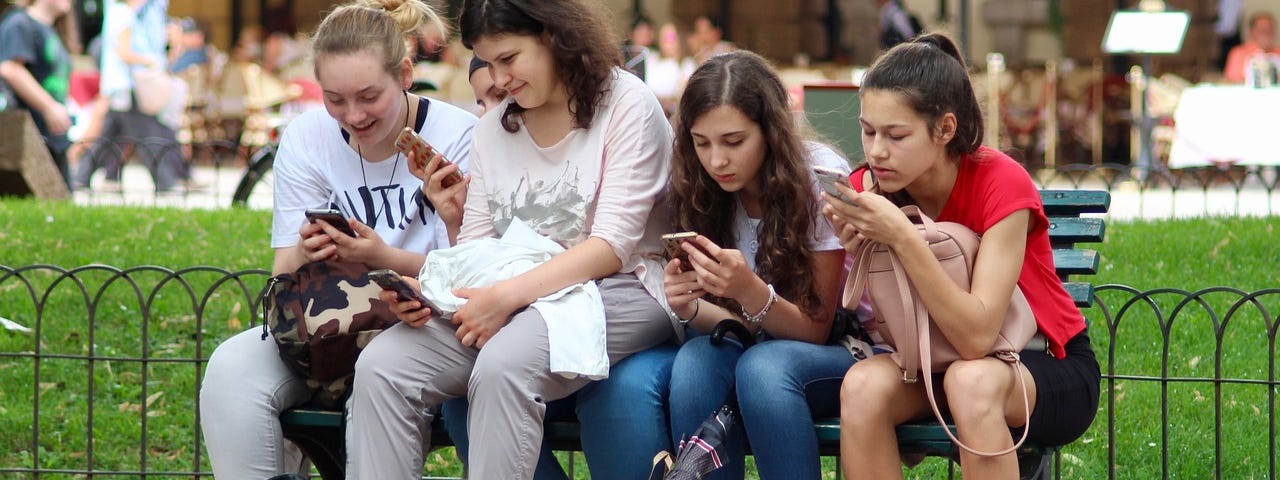 IMAGE: Four girls sitting on a bench and checking their smartphones