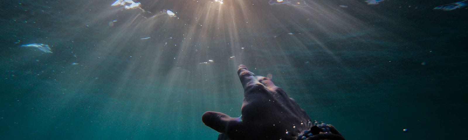 Someone underwater reaching towards the surface of the water through which sunlight reaches out towards them.