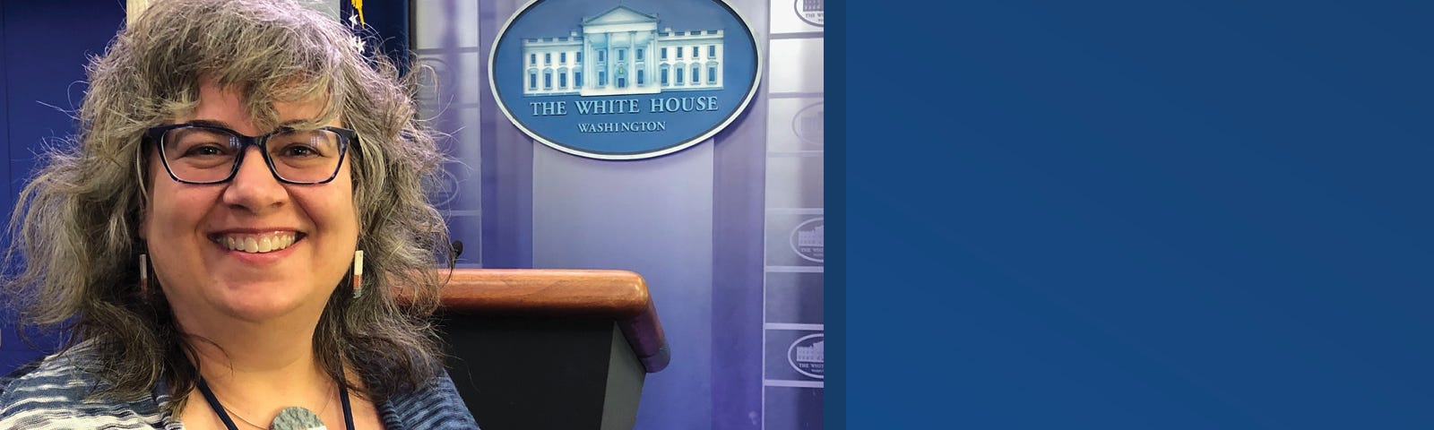 A woman stands in front of a wooden podium and the seal of The White House. She has shoulder-length grey and black hair and wears black glasses. There is a vertical blue line and to the right of that line is white text on a blue background that reads “Martha Wilkes, designer & accessibility strategist”