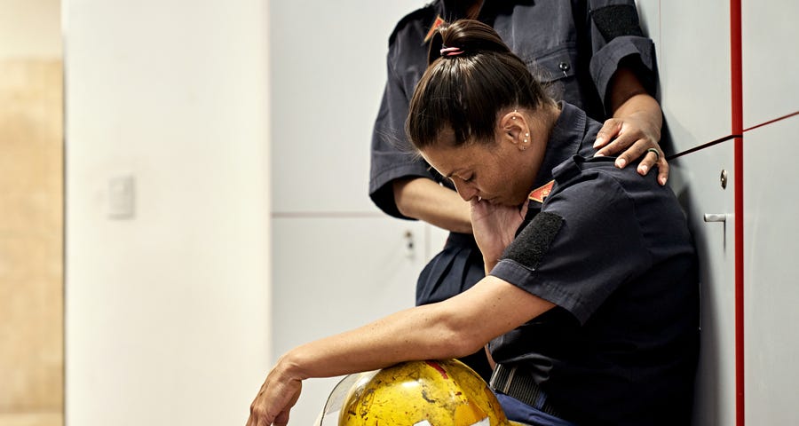 A woman in uniform sitting in a locker room with helmet in lap, head down, and eyes closed, being consoled by a colleague.