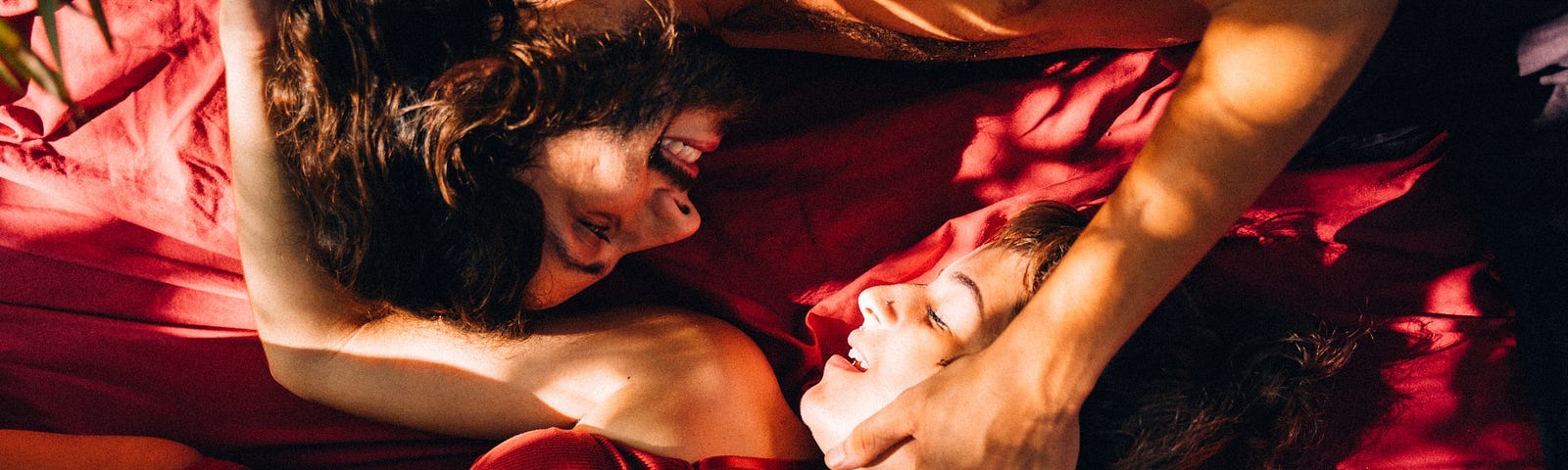 man and woman in bed with red lingerie and red sheets, smiling & in love.