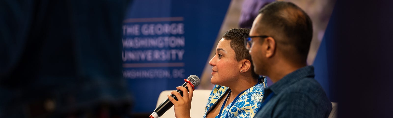 A woman with short brown hair wearing a blue and yellow sundress holds a microphone while sitting in white chairs next to a man with short hair and a blue shirt on a stage at a conference.