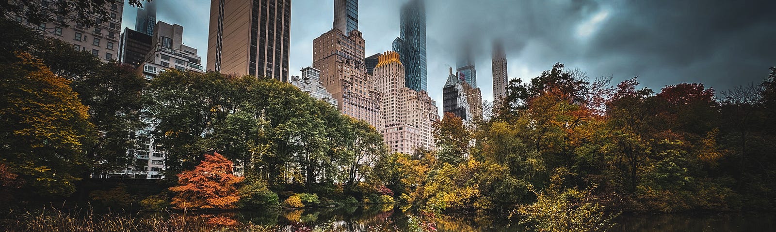 NYC skyscrapers with the tops of the buildings obscured by dark clouds with a view of fall foliage in Central Park in the foreground.