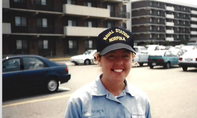 Young woman with Naval Station Norfolk ballcap on and a Navy blueshirt on, standing in a parking lot, with a large government building behind her; photo is from the chest up.