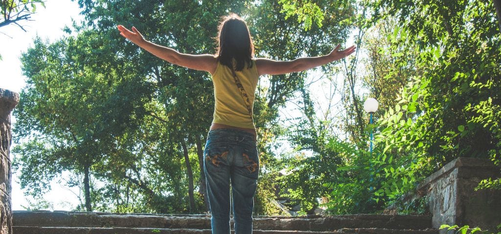 Woman with Arms outstretched, palms upward facing towards the sky an eyes raised looking into the trees. She is saying — Thank You God for this beautiful day.