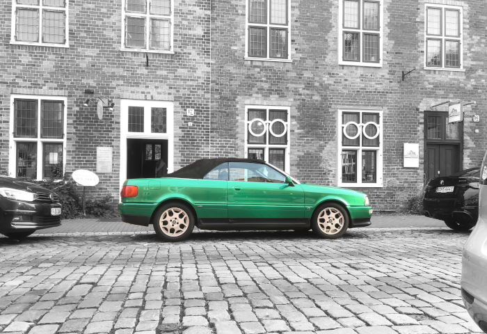 Metallic green convertible soft top roof car with decolourised background of cobblestone road with ancient building in background in Potsdam, Germany.