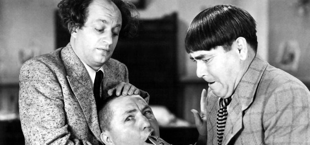 Black and white photo of the Three Stooges showing Curly Joe having his tooth pulled with a pair of pliers by Moe, while Larry holds Curley’s head still.