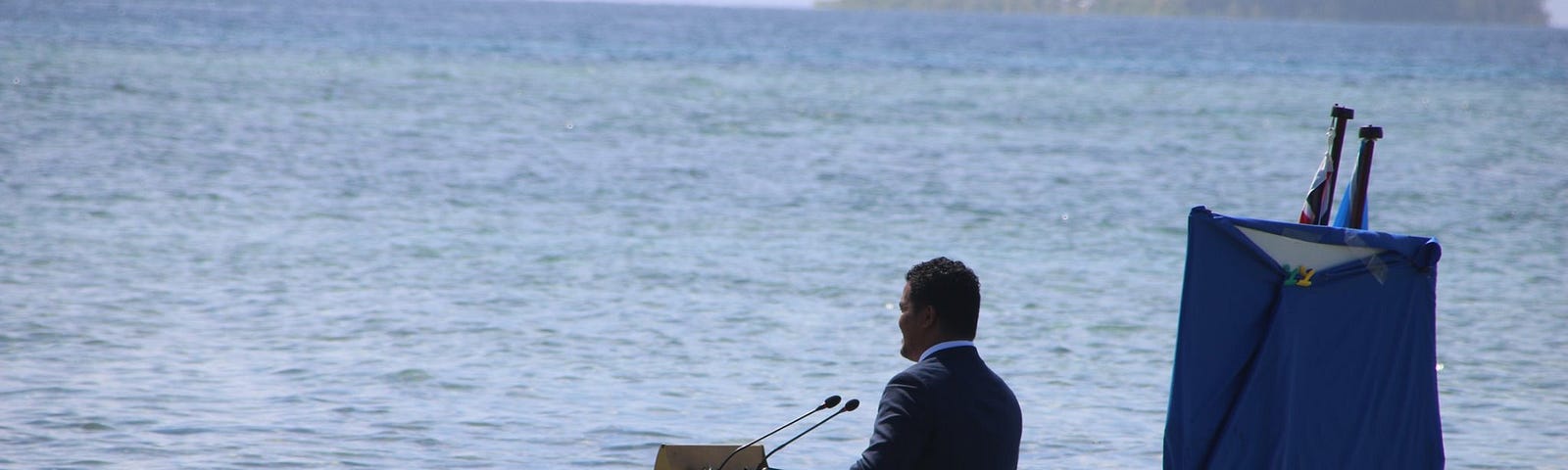 A man is standing knee-deep in water. In front of him is a desk with two microphones, behind him two flags.