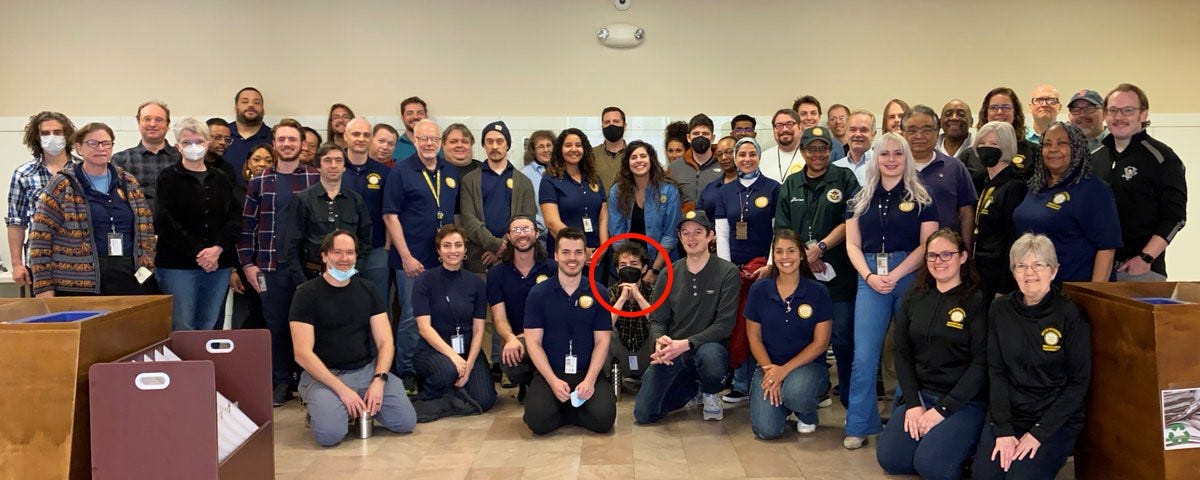 A group photo of Pittsburgh’s Department of Innovation and Performance, taken in an office lobby. Many employees wear garments with the I&P logo. A red circle marks the author crouching near the front masked.