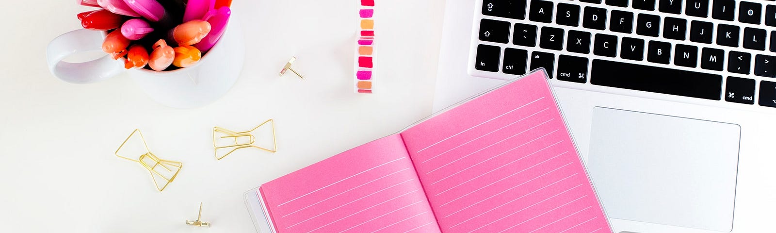 A silver laptop is. open on a white desk. Laying on the desk is a pink notebook, a container with pencils in shades of pink and gold, paper clips and reels of thin decorative tape.