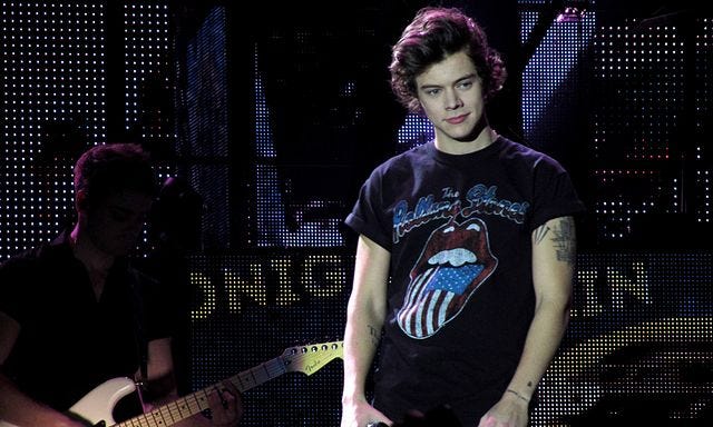 Harry wearing Rolling Stones T shirt, hands in pockets, in front of performance screen, bassist in background