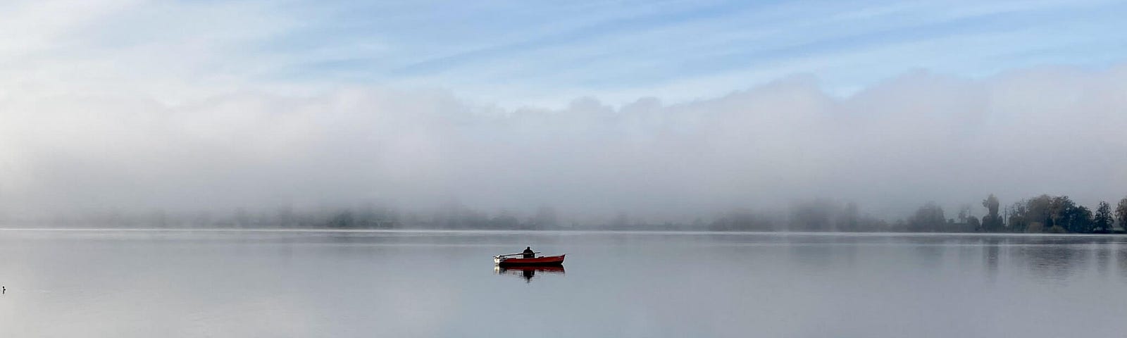 A small rowboat with a solitary fisherman on a large, smooth lake surface