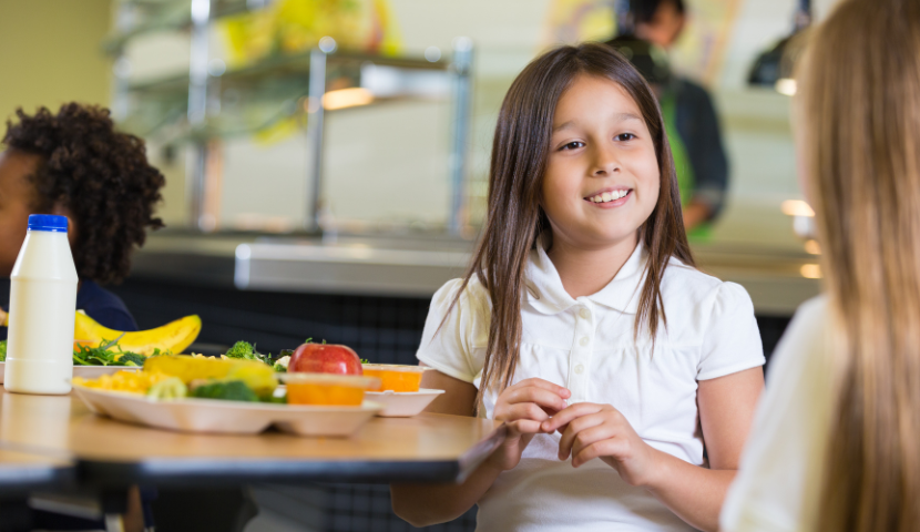 A student sits at a table with a school meal on it, talking to a friend (off-screen)