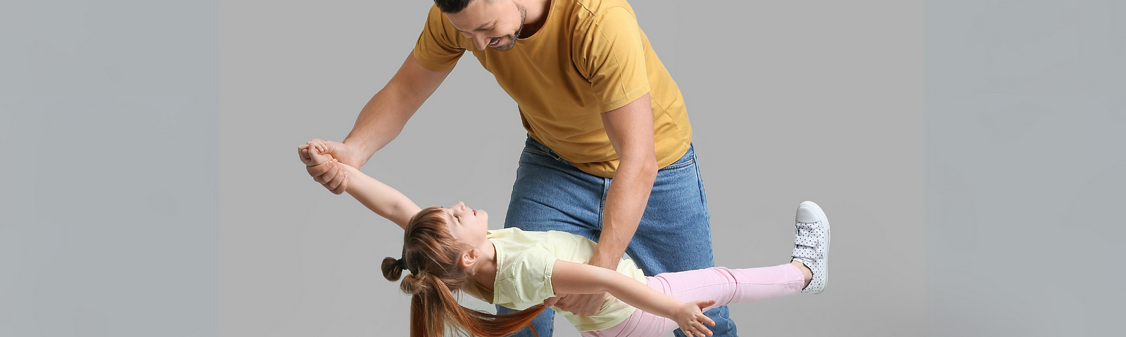 This image shows a man and a young girl in the midst of a playful dance or twirl. The man is bending slightly, holding the girl’s hand and arm as she leans back with her other arm extended, her foot kicking up behind her.
