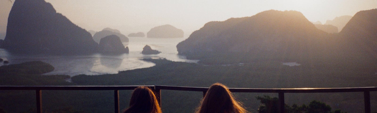 Two girls on a cliff look out toward the ocean sunset.