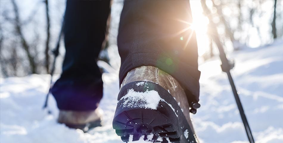 A view from the ground of a pair of legs adorned with winter hiking boots moving through the snow.